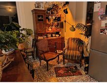Angolo cucina-soggiorno con mobili in legno massello e credenza piena di stoviglie, lampade da terra e poltrone imbottite. Pavimento con piastrelle decorative a motivi geometrici, molte piante in vaso e frigorifero a vista sulla destra.