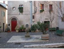 Piccola piazza lastricata in ciottoli con una fontanella in pietra al centro, piante in vaso e panchine verdi ai lati. Sullo sfondo, facciate di edifici in intonaco grezzo con porta ad arco rossa, finestre con persiane e una lanterna a parete.