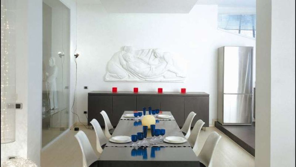 Sala da pranzo moderna in un open space luminoso, con tavolo rettangolare scuro apparecchiato e sedie bianche. Sullo sfondo un mobile basso con candele rosse e un grande bassorilievo bianco a parete, accanto a un frigorifero in acciaio inox e una parete in vetro.