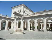 Cortile interno in pietra chiara con porticati ad arcate su colonne classiche e cornici decorative lungo il perimetro. Al centro si trova un pozzo in muratura, con un edificio a più piani sullo sfondo sotto un cielo sereno.
