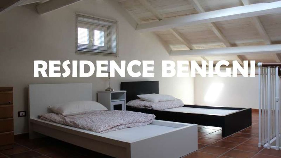 Camera da letto mansardata con soffitto in legno a travi bianche, pavimento in cotto e due letti bassi con comodino; una finestra con veneziane illumina l’ambiente vicino a una ringhiera interna.
