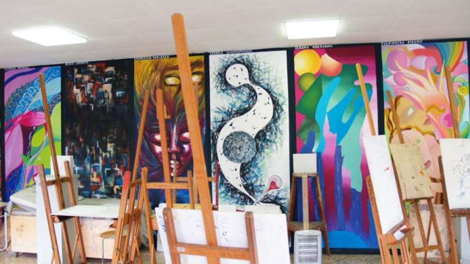 Aula di arte con cavalletti in legno e tele appoggiate, illuminata da pannelli a soffitto. La parete di fondo è coperta da grandi murales colorati e astratti.