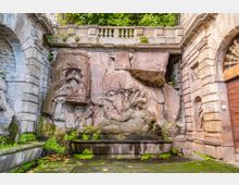 Cortile in pietra di un edificio storico con una grande fontana addossata al muro, decorata da bassorilievi scolpiti nella roccia e vasca rettangolare con muschio. Intorno si vedono pareti in blocchi di pietra, un arco e una balaustra superiore con vegetazione.