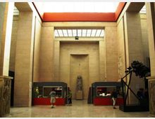 Ampio atrio interno di un museo con alte pareti in pietra chiara, soffitto a lucernario e fascia superiore rossa. Nella sala si vedono vetrine espositive rosse ai lati, una statua su piedistallo al centro e un grande oggetto scultoreo scuro sulla destra.