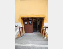 Scala esterna in pietra che scende a un ingresso seminterrato incassato in una parete gialla, con corrimano metallici ai lati. In fondo si vede una porta marrone con locandine e una porta vetrata scorrevole.