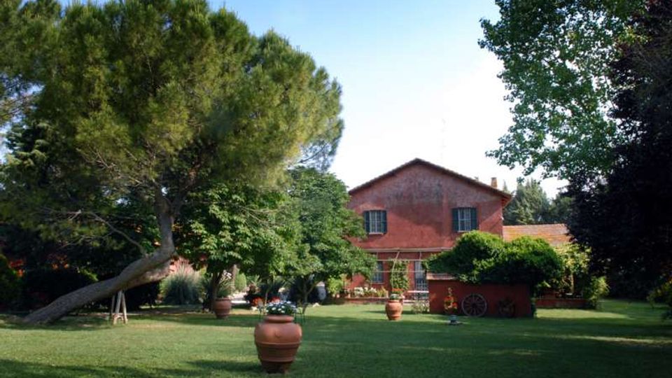 Ampio giardino con prato curato, grandi alberi e vasi in terracotta distribuiti sul terreno. Sullo sfondo si vede una casa rurale in mattoni rossi con finestre e pergolato, circondata dalla vegetazione.