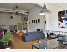 Ampio loft open space con pavimento in legno, soffitto alto con travi e grandi lampade industriali a sospensione. Zona soggiorno con divano grigio e pouf, tavolo in vetro e scaffali con libri e dischi lungo le pareti bianche decorate con quadri.