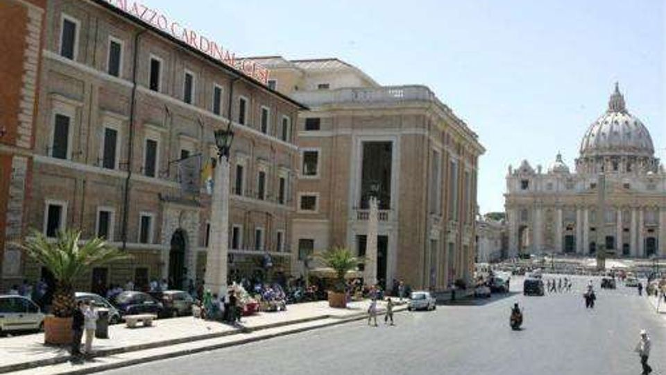 Ampia piazza urbana con strada asfaltata e palazzi storici a più piani con facciate in pietra e finestre regolari; sullo sfondo si vede la Basilica di San Pietro con la grande cupola. Ai lati sono presenti lampioni, alcune palme in vaso e auto parcheggiate.