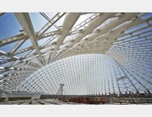 Interno di una grande struttura architettonica moderna con copertura a griglia di travi metalliche bianche e pannelli trasparenti, che forma una volta curva. Ampie aperture lasciano vedere il cielo e, sullo sfondo, una sagoma urbana con gru e edifici.