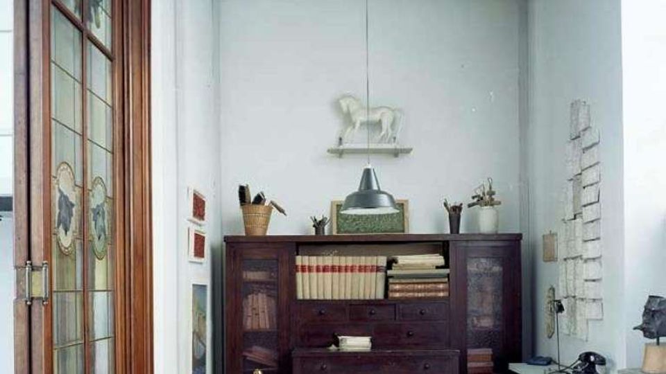 Piccolo studio interno con pareti bianche, grande armadio in legno scuro pieno di libri e oggetti e un tavolo bianco al centro illuminato da una lampada a sospensione. A sinistra c’è una porta in legno con vetri decorati, mentre a destra si vedono una parete con fogli appesi e un termosifone con telefono e sgabello.