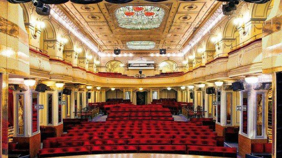 Interno di un teatro storico con platea di poltrone rosse disposte in file e due ordini di balconate laterali con colonne e lampade. Sul soffitto decorato si vedono riquadri ornamentali e un grande lucernario centrale, con un ampio sipario scuro in alto.