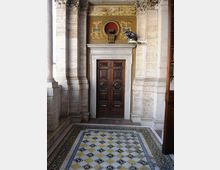Ingresso interno di un edificio storico con grandi colonne in pietra e una porta doppia in legno scuro incorniciata da una cornice chiara. In primo piano si vede un pavimento a mosaico geometrico con motivi a rombi e bordure decorative.