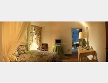 Camera da letto in stile rustico con letto a baldacchino e tessuti floreali, pareti bianche e pavimento in cotto. Un tavolino con lampada e telefono, una piccola TV su tavolo e un passaggio ad arco che conduce a un bagno con piastrelle decorative.