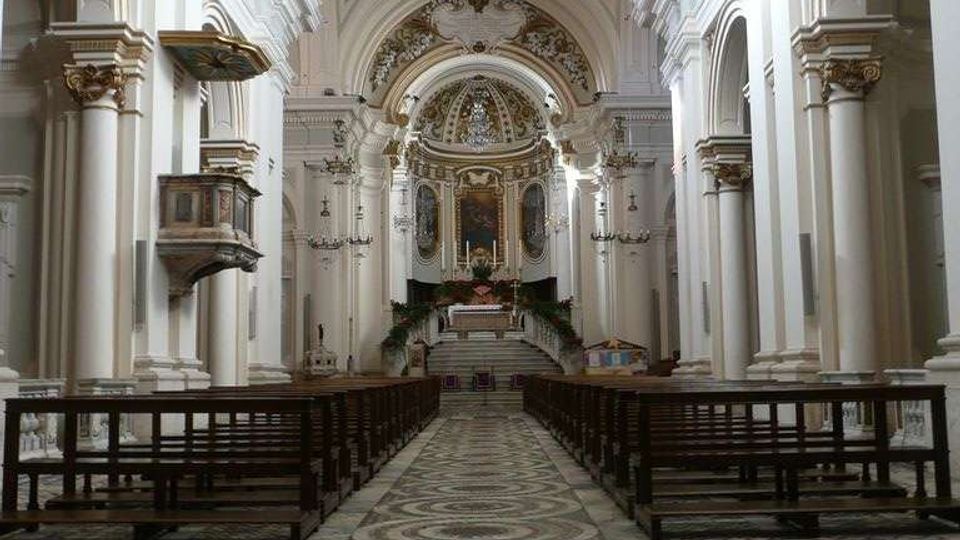 Interno di una chiesa con navata centrale lunga, pavimento a mosaico con motivi circolari e file di panche in legno ai lati. Alti archi e colonne bianche sorreggono volte decorate, con altare rialzato e cupola ornata sullo sfondo e una finestra circolare in alto.