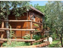 Casetta in legno con tetto spiovente, circondata da alberi e vegetazione, con staccionata in tronchi e piccola aiuola rialzata in mattoni. Sulla facciata sono visibili una lanterna a parete e una catena pluviale.