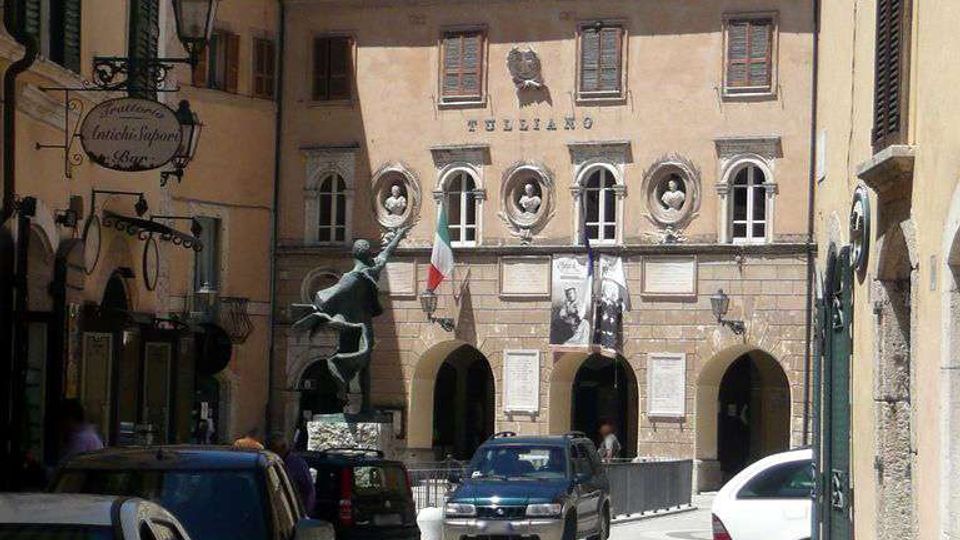Via del centro storico che conduce a una piazzetta con un palazzo in stile classico, facciata chiara con portici ad archi e finestre ovali decorative. Lampioni in ferro alle pareti, bandiera italiana e striscioni sulla facciata, con auto parcheggiate lungo la strada.