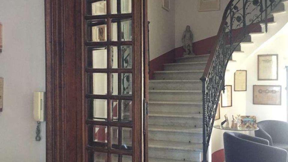 Ingresso interno di un edificio con grande porta ad arco in legno e vetri, che si apre su una scalinata in marmo curvilinea con ringhiera in ferro battuto. Sulle pareti bianche con fascia rossa sono appesi quadri e cornici, con una piccola area salotto con poltrone scure.