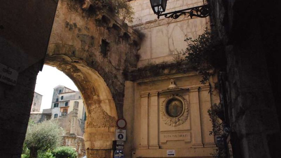 Antico passaggio ad arco in pietra che collega edifici in muratura, con una facciata decorata da un’edicola con medaglione incassato e cornici scolpite. In alto si vede un lampione in ferro battuto e, sullo sfondo, una piccola piazza con vegetazione e costruzioni storiche.