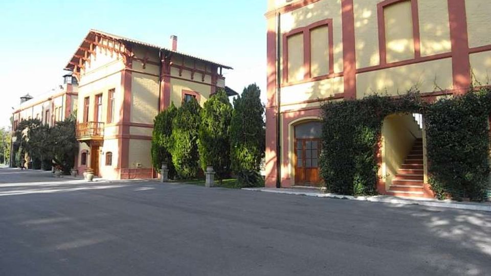 Complesso di edifici in stile villa storica con facciate color crema e cornici rosse, affacciati su un ampio piazzale asfaltato. Sono visibili finestre alte, un portico con scala d’ingresso e siepi e alberi sempreverdi lungo i lati.