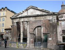 Facciata di un edificio storico in muratura e pietra, con frontone triangolare, grandi archi murati e colonne antiche parzialmente conservate. Davanti si vede una piazza con transenne metalliche e, sullo sfondo, palazzi adiacenti e una cupola visibile a destra.