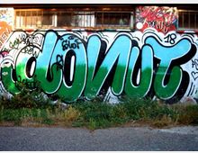 Muro esterno di un edificio con finestre protette da grate, ricoperto da un grande graffito con lettere blu e verdi su sfondo bianco. In primo piano si vede una striscia di asfalto con erba e vegetazione bassa lungo la base del muro.
