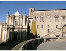 Facciata di una chiesa barocca in pietra chiara con colonne e timpano, accanto a un grande edificio storico in muratura con finestre ad arco e campanile. In primo piano si vede una terrazza o ponte in pietra con balaustra curva e una rampa di scale sul lato destro, sotto cielo sereno.