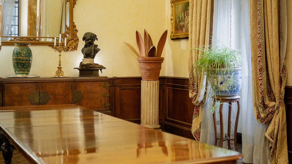 Sala da pranzo in stile classico con lungo tavolo in legno su tappeto orientale rosso, boiserie scura e grande specchio dorato sopra una credenza. Ampia finestra con tende pesanti e velo bianco, con pianta su piedistallo, soffitto alto decorato e arredi d’epoca.