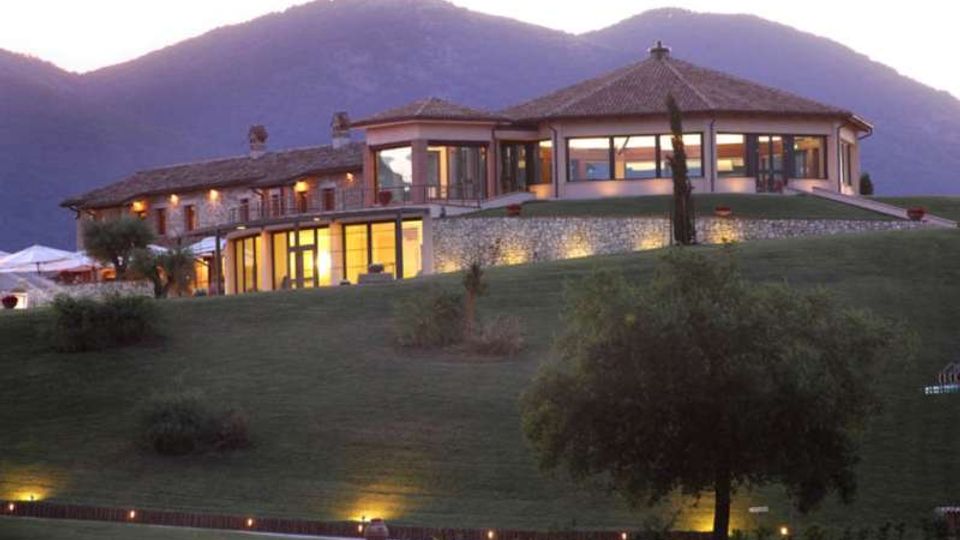 Edificio tipo resort o agriturismo su una collina erbosa, con tetti in coppi e grandi vetrate illuminate. Sullo sfondo si vedono montagne al crepuscolo e, in primo piano, alberi e un vialetto con luci a terra.