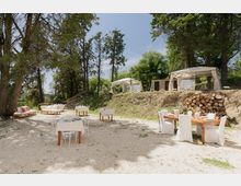 Area esterna attrezzata per ristorazione in un bosco, con tavoli su ghiaia e sedie con coperture bianche sotto alberi alti. Su un terrapieno sullo sfondo ci sono due gazebo bianchi e una catasta di legna accanto a una parete in pietra.