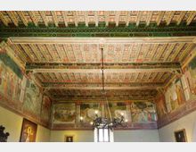 Interno di una sala storica con soffitto a cassettoni dipinto e travi decorate, e pareti affrescate con scene figurative lungo il perimetro superiore. Al centro pende un grande lampadario in ferro battuto sopra una finestra e quadri appesi alle pareti.