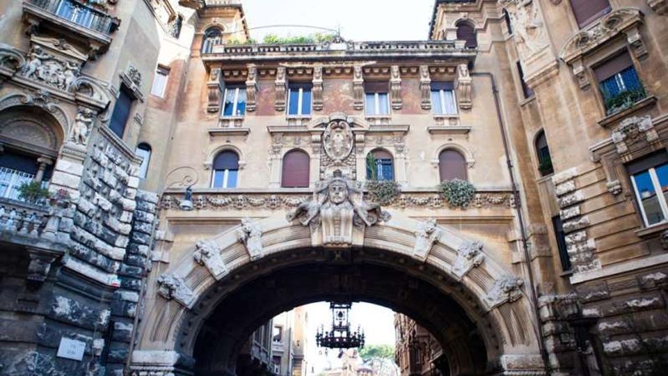 Arco monumentale in pietra che collega due edifici storici, con facciata decorata da bassorilievi e stemmi scolpiti al centro. Sotto la grande volta si intravede una strada urbana e un lampadario sospeso.