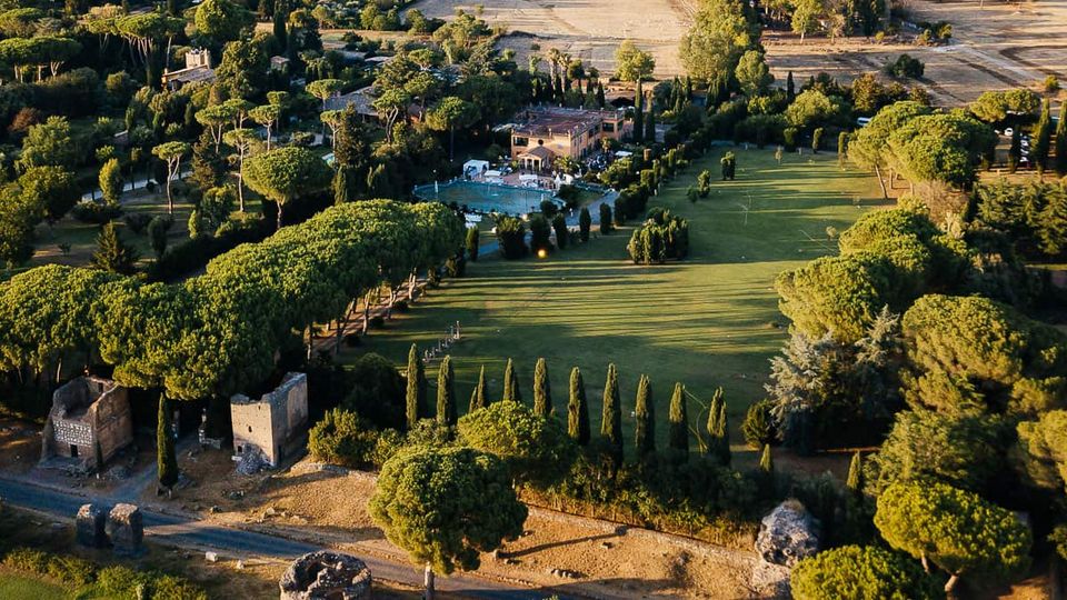 Veduta aerea di una tenuta di campagna con ampio prato centrale, filari di cipressi e boschi di pini mediterranei, con alcuni edifici in pietra e una piscina vicino alla villa principale. Ai margini si vedono strade sterrate e campi coltivati.