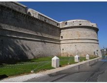 Mura esterne di una fortezza in pietra chiara con grande bastione cilindrico e cornici orizzontali. Davanti si vede una fascia di prato delimitata da paletti con cavi e una strada asfaltata lungo il perimetro sotto un cielo sereno.