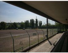 Tribuna coperta di un impianto sportivo con gradinate in cemento, affacciata su un campo in terra battuta recintato con rete metallica. Sullo sfondo si vedono alberi, pali dell’illuminazione e una piccola porta da calcio.