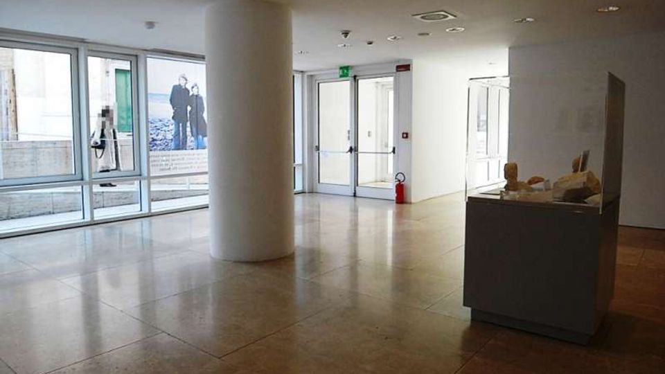 Atrio interno moderno e luminoso con pavimento in pietra lucida, grandi finestre e porte a vetri, e una colonna cilindrica bianca al centro. Sulla destra è presente una teca espositiva su un basamento, con un estintore vicino a una porta di uscita.