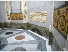 Interno decorato con pareti rivestite in marmo venato e pannelli ornamentali gialli con motivi a volute. Pavimento geometrico in marmo con riquadri e cerchi colorati, delimitato da zoccolature in pietra scura.