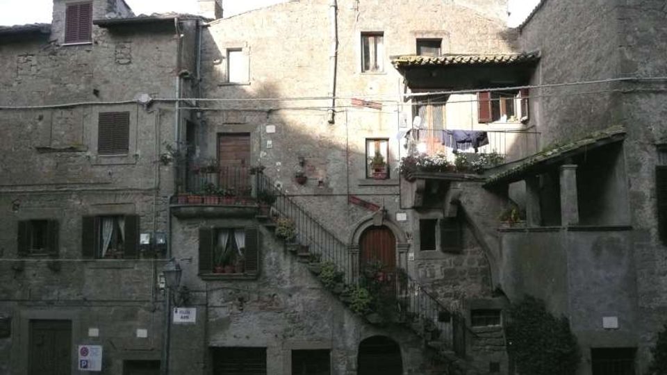 Facciata di un complesso di case in pietra in un borgo storico, con finestre e portoni scuri, balconi con vasi e una scalinata esterna in muratura con ringhiera. Un piccolo loggiato coperto e tetti in coppi completano l’insieme delle costruzioni addossate tra loro.