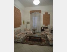 Salotto interno con divani e poltrone imbottiti a fantasia floreale disposti attorno a un tavolino rettangolare con piano in vetro su tappeto decorato. Sulla parete di fondo c’è una grande portafinestra con tende leggere, con lampade e mobili d’appoggio ai lati e pannelli decorativi alle pareti.