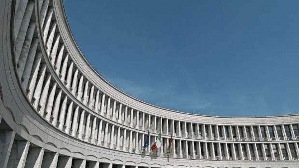 Facciata curva di un grande edificio istituzionale in cemento e vetro, con file regolari di finestre e pilastri verticali su più piani. Al centro sono visibili aste con bandiere davanti a un ampio spazio pavimentato sotto un cielo azzurro.