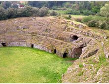 Anfiteatro antico scavato nella roccia, con gradinate semicircolari in pietra e piccole aperture ad arco lungo il perimetro. Al centro si vede un’area erbosa, circondata da vegetazione e colline sullo sfondo.