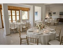Sala ricevimenti luminosa con grandi finestre a telaio in legno e colonne bianche, allestita con tavoli rotondi coperti da tovaglie bianche. Sedie dorate, centrotavola floreali e candele completano l’apparecchiatura, con quadri e lampade sulle pareti sullo sfondo.