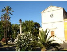 Facciata di una piccola chiesa intonacata di bianco con rosone circolare e croce sul frontone, affacciata su una piazza. In primo piano ci sono palme, un cespuglio fiorito e un lampione, con alberi sullo sfondo sotto un cielo sereno.