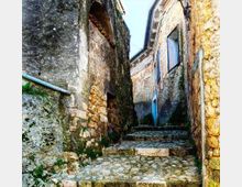 Vicolo stretto in pietra con pavimentazione acciottolata e scalini che salgono tra antiche case in muratura. Le pareti in pietra grezza mostrano segni di umidità e muschio, con finestre e una grondaia visibili lungo la salita.