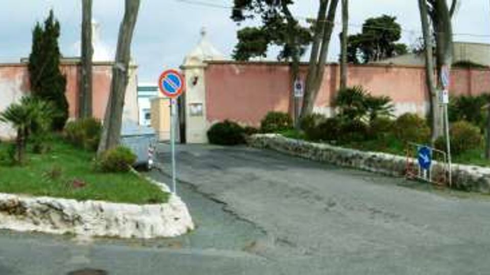 Ingresso carrabile a un complesso recintato da un lungo muro rosa con portale centrale, aiuole con pietre bianche e vegetazione mediterranea. Strada asfaltata che si biforca ai lati, con pini alti, segnaletica stradale e un edificio basso sullo sfondo.