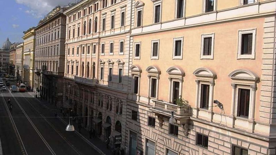 Veduta in prospettiva di una lunga strada urbana fiancheggiata da alti palazzi storici dai toni chiari, con facciate ornate da cornici e finestre regolari. La carreggiata è ampia e quasi vuota, con lampioni sospesi e cielo parzialmente nuvoloso sullo sfondo.