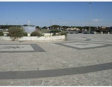 Ampia piazza pavimentata in pietra con grandi riquadri decorativi sul suolo e una fontana circolare al centro, circondata da aiuole. Sullo sfondo si vedono una strada con auto, lampioni alti e una linea di alberi sotto un cielo sereno.