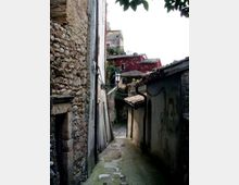 Vicolo stretto in un borgo antico, con pavimentazione in pietra e muri alti in pietra e intonaco ai lati. Una lanterna a parete e cavi sospesi attraversano il passaggio, con case dai tetti in tegole visibili sullo sfondo in salita.