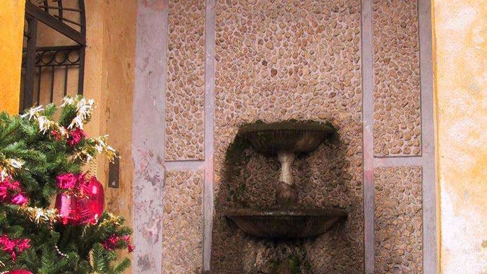Cortile con pareti intonacate e nicchia ad arco rivestita in pietre, con fontana in pietra a due vasche e vasca inferiore semicircolare. A sinistra si vede una finestra ad arco con inferriata e un albero decorato con grandi ornamenti rossi.