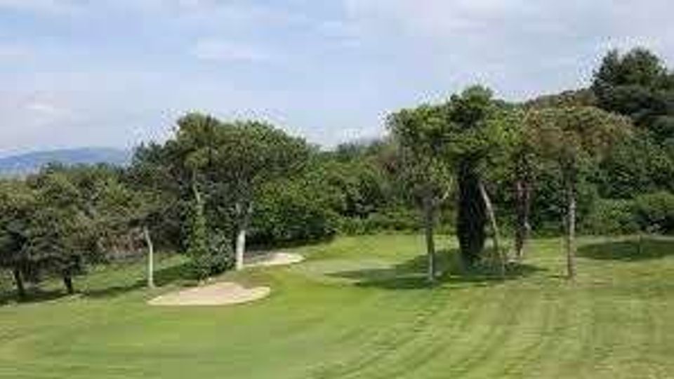 Campo da golf con ampio prato verde curato, un bunker di sabbia e alberi alti che delimitano il fairway. Sullo sfondo si vedono colline e un cielo parzialmente nuvoloso.