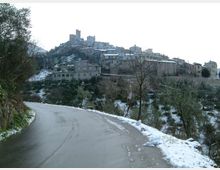 Strada asfaltata in curva con bordi innevati che conduce verso un borgo medievale arroccato su una collina, con case in pietra e torri in cima. Vegetazione di alberi e pendii con chiazze di neve circondano l’abitato.